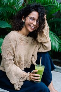 A woman with curly hair, wearing glasses, enjoys a healthy green smoothie surrounded by lush plants.